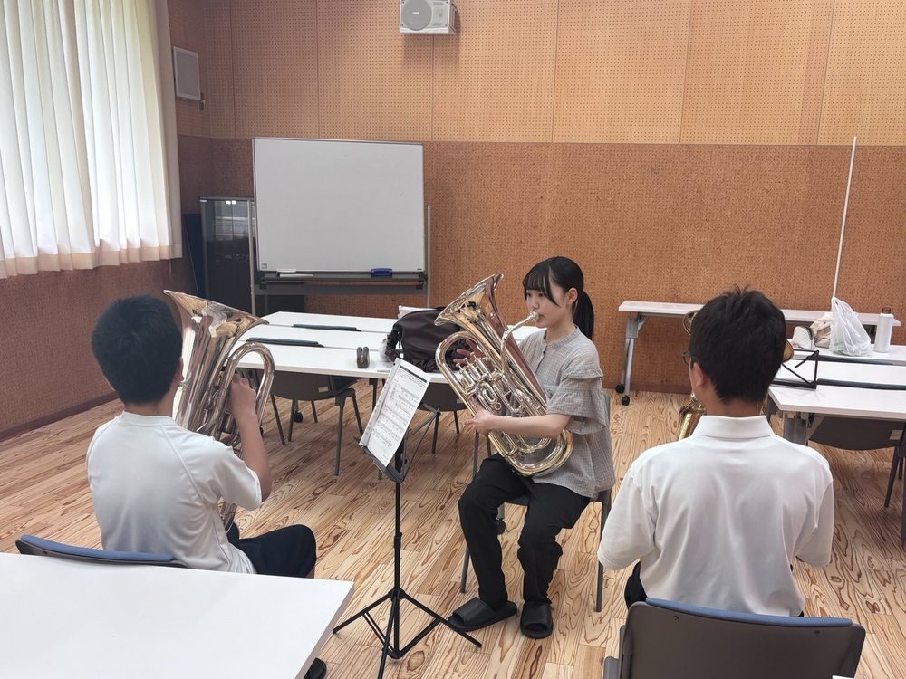 大学生がユーフォニアムを吹いて、中学生に低音楽器を指導している様子